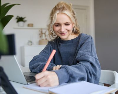 Portrait of young woman, lifestyle blogger, recording video of herself, making notes, writing in journal, sitting in front of laptop in a room and studying
